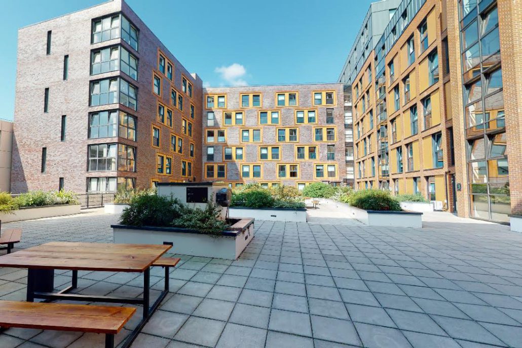 A courtyard surrounded by modern apartment buildings with large windows, featuring benches, planters, and a clear blue sky. No people are visible.