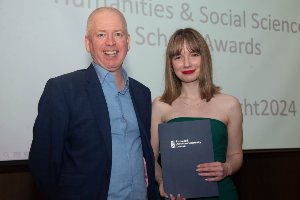 Two people are smiling in a room with a "Humanities & Social Sciences Awards" sign. One holds an award from ϲֱֳ American University London.