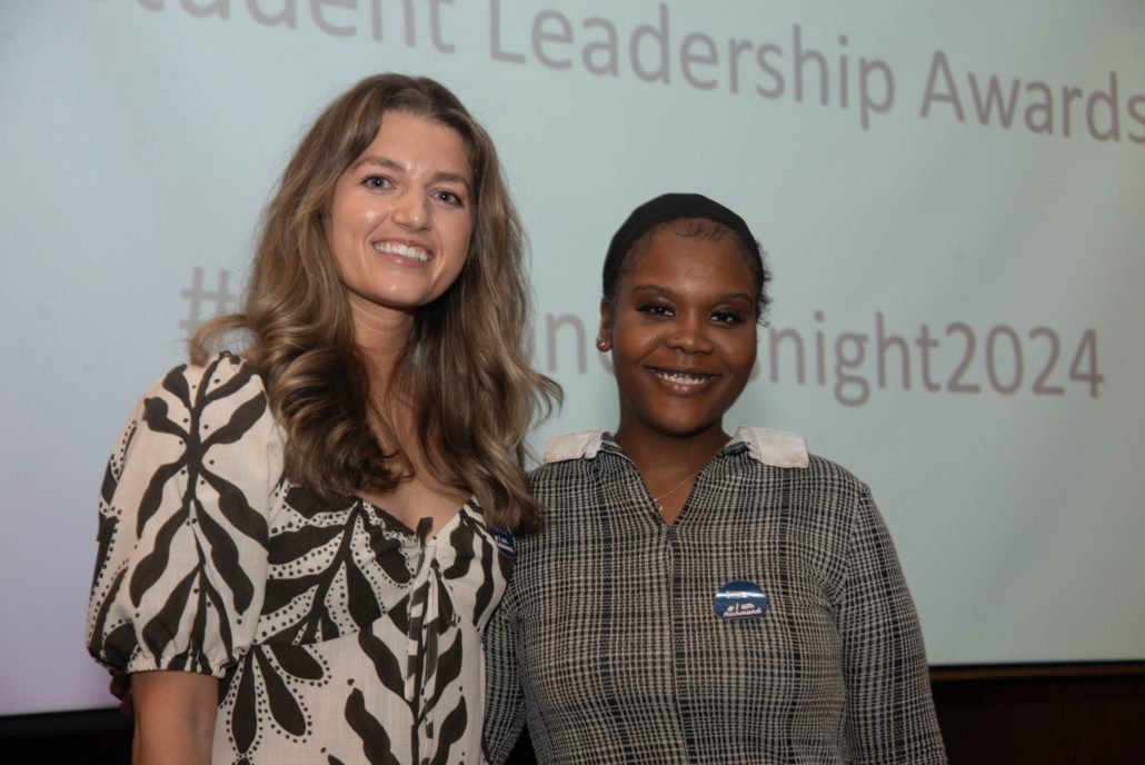 Two smiling persons pose in front of a screen displaying "Student Leadership Awards" and a hashtag with the year 2024, suggesting an awards event setting.
