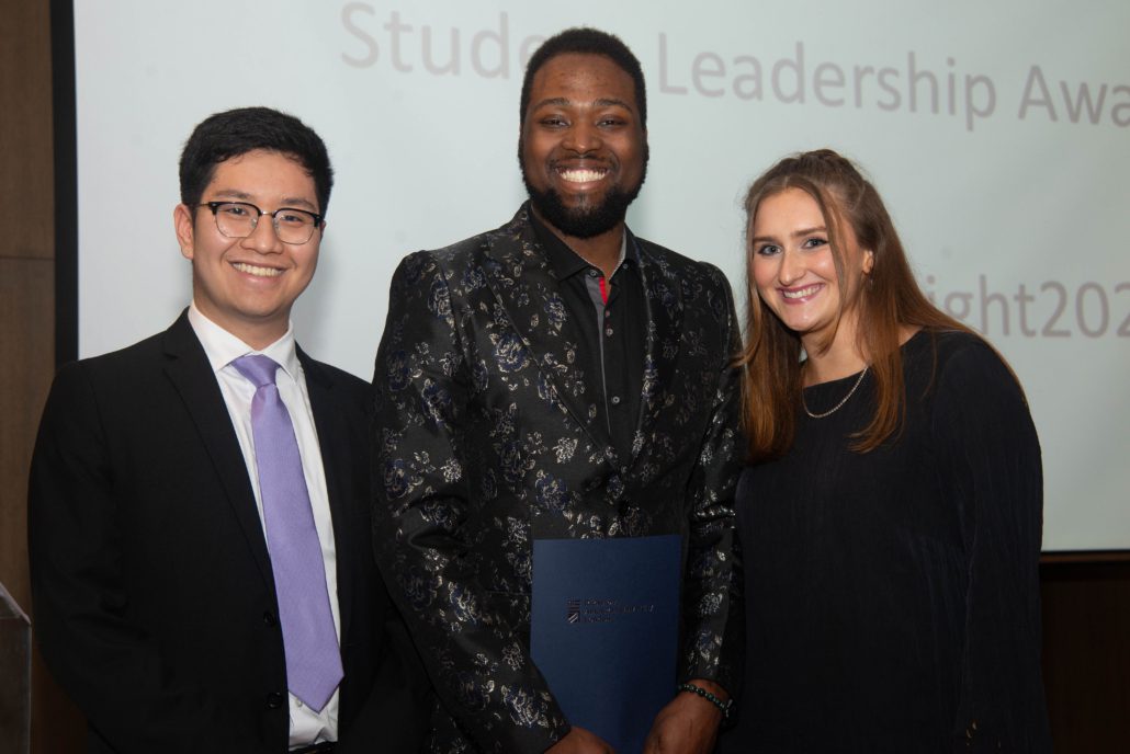 Three smiling people stand together, two holding a certificate, at a "Student Leadership Awards" event. They're dressed formally, suggesting a celebratory occasion.
