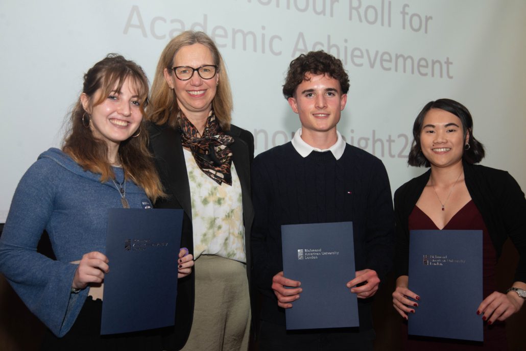Four individuals smiling, holding certificates, in front of a screen with text "Honour Roll for Academic Achievement". They appear happy and accomplished.