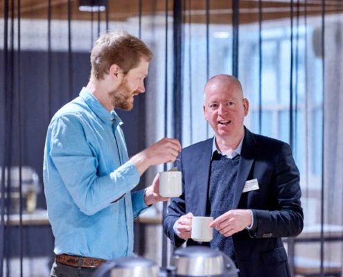 Two people are holding mugs and conversing in a modern indoor setting with blurred background. One person gestures while talking to the other, who listens attentively.