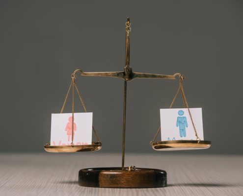 A traditional balance scale with two drawings representing people, one pink and one blue, suggesting a concept of gender equality or comparison.