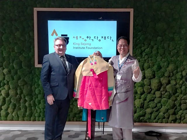 Two people stand smiling beside a traditional Korean hanbok on a mannequin, with a sign for the King Sejong Institute Foundation in the background.