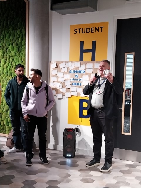 Three people are standing in a modern room with decorative greenery, a sign reading "STUDENT", and sticky notes on a wall. One person speaks into a microphone.