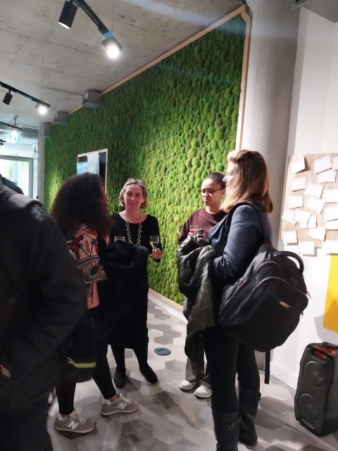 A group of individuals is having a conversation in an indoor space, with one person holding a glass. A green textured wall is featured behind them.