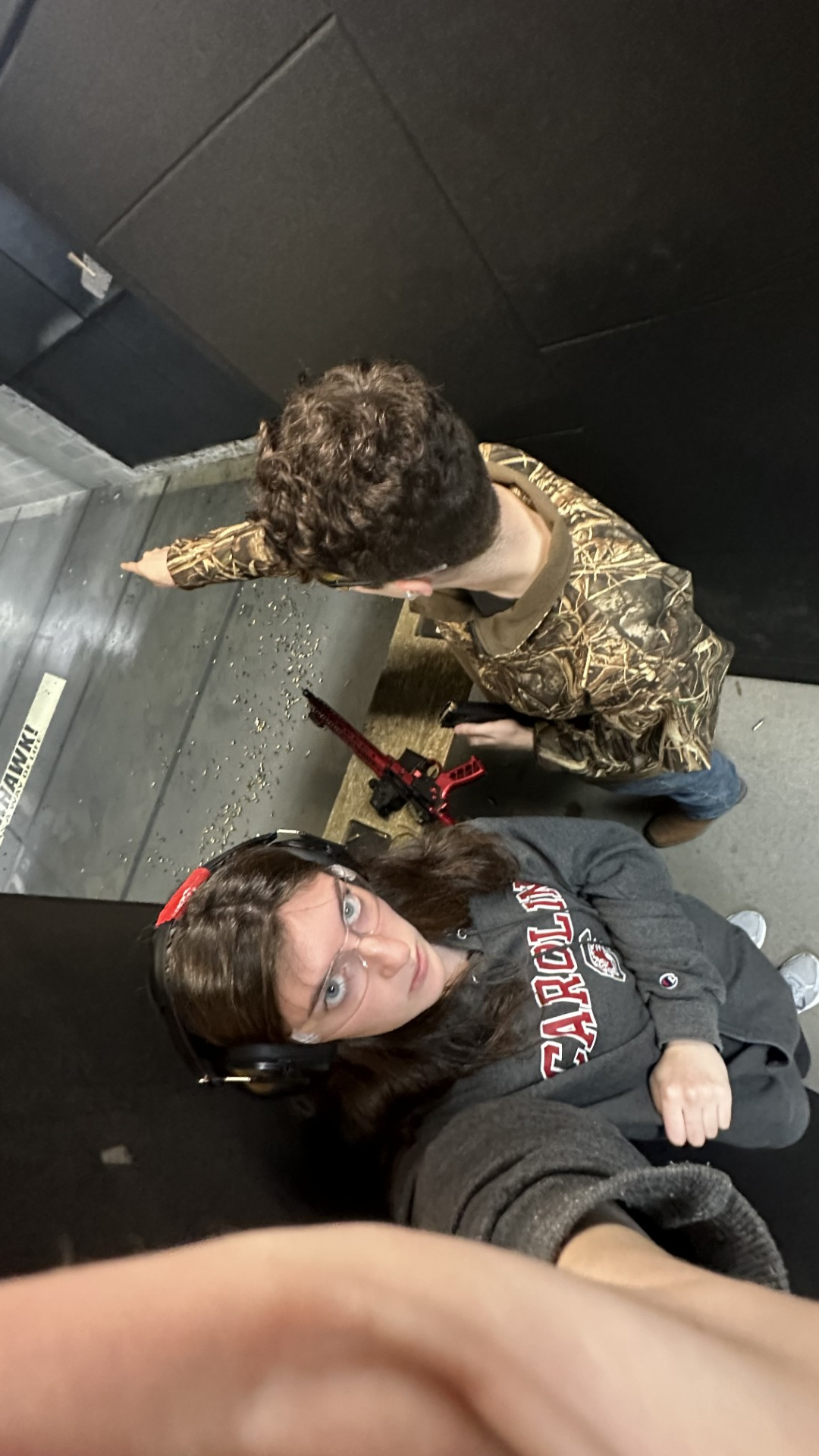 The image shows two people at a shooting range. The person in the foreground is wearing protective headphones and eyewear, while the other is aiming a rifle.