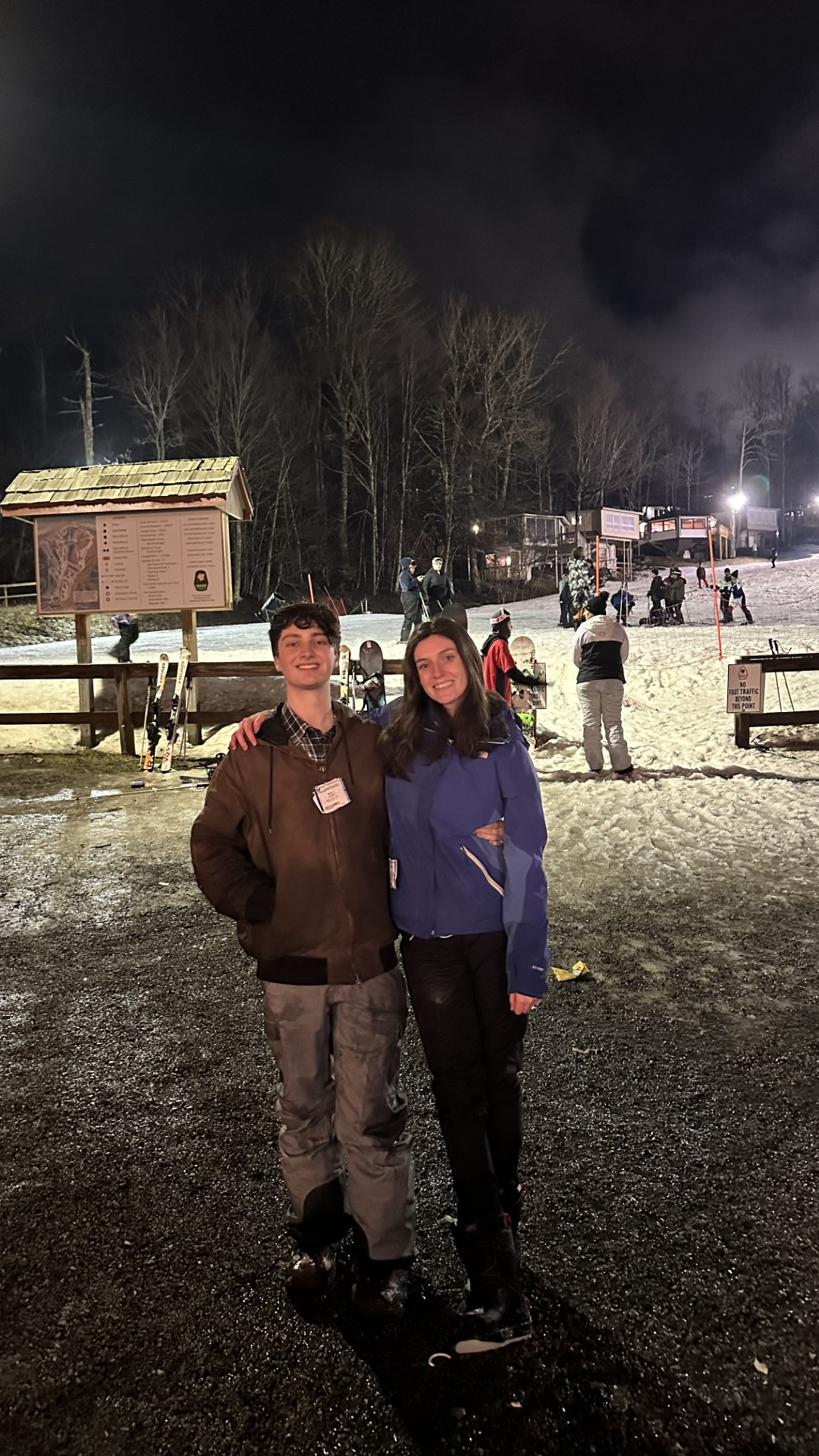 Two people are smiling in the foreground at a snowy night-time ski resort, with skiers, a lift, and a well-lit lodge in the background.