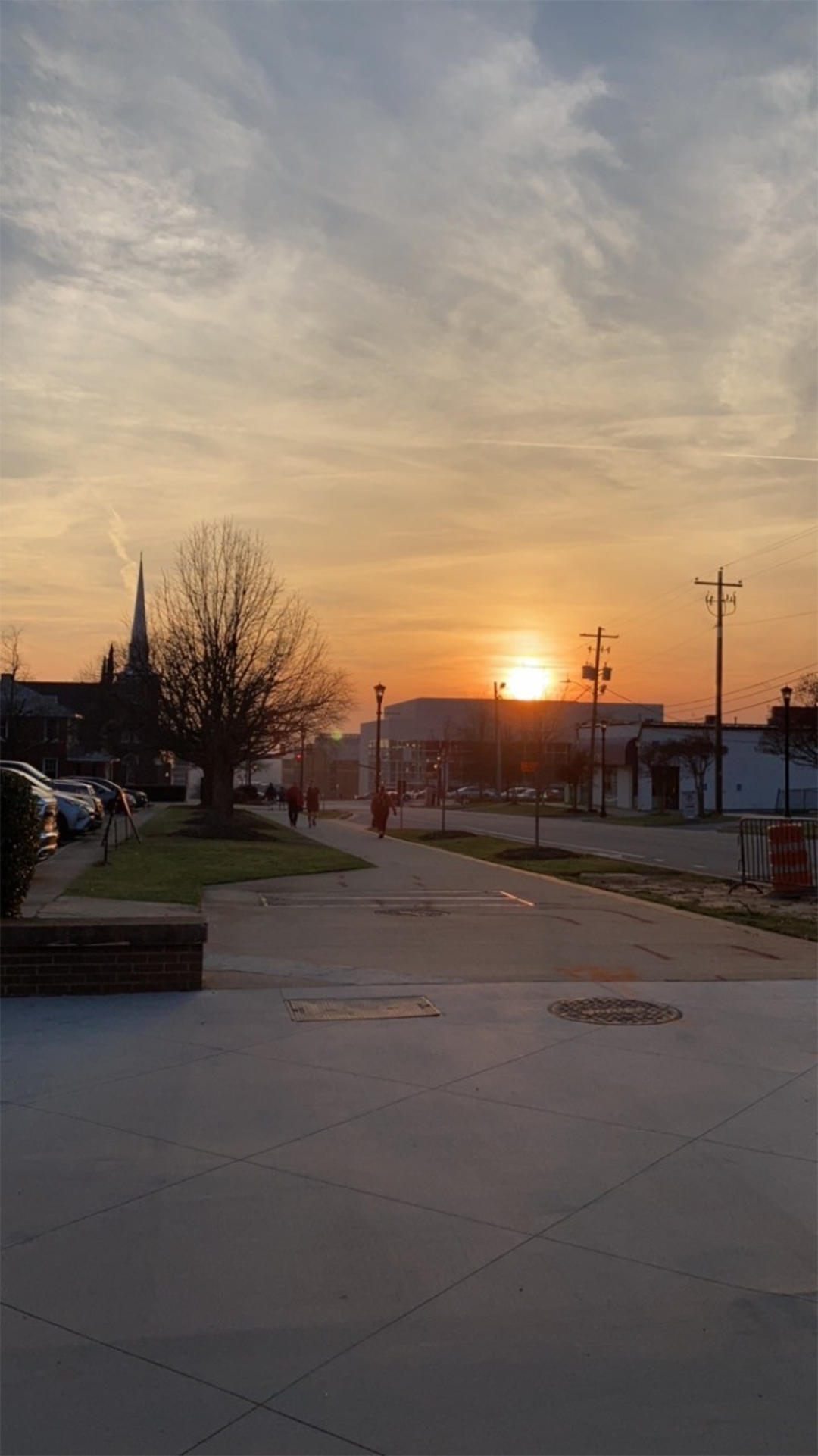 The image captures a serene sunset with orange hues behind a distant steeple, overlooking a quiet urban streetscape with people walking and cars parked.
